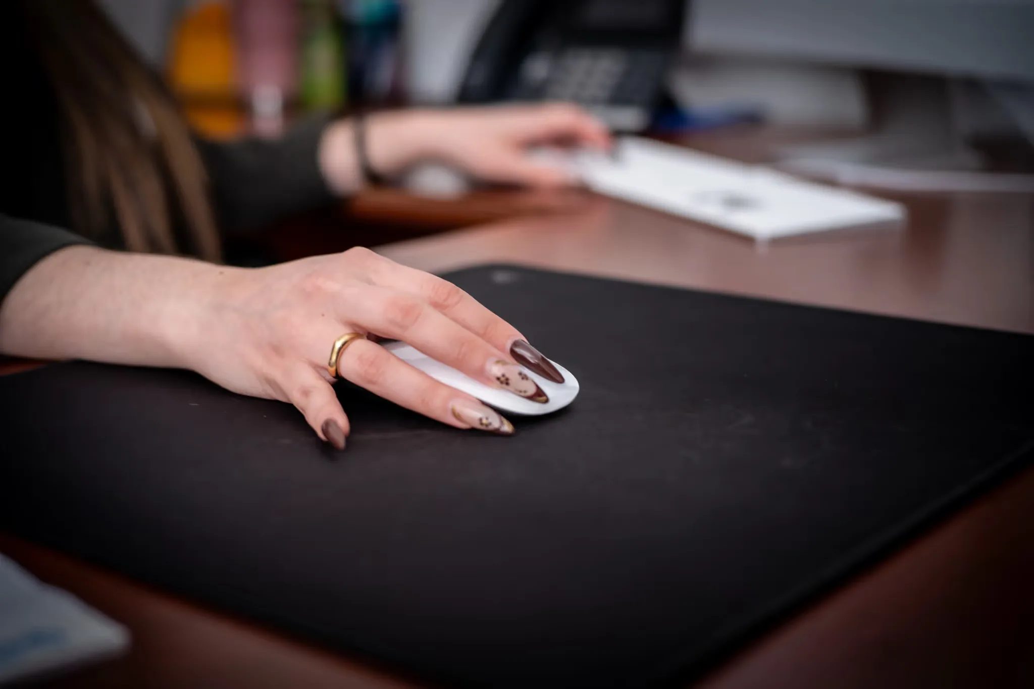 Close-up of a hand with manicured nails on a white mouse on a black desk mat, keyboard and desk phone blurred behind.