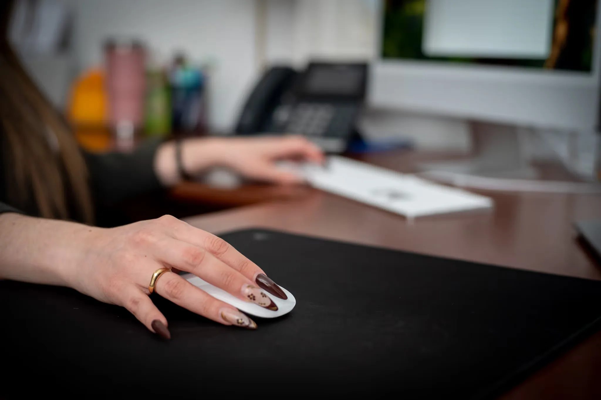 Close-up of a hand on a white mouse on a black desk mat at Nelson Law Firm, an iMac and desk phone blurred behind.
