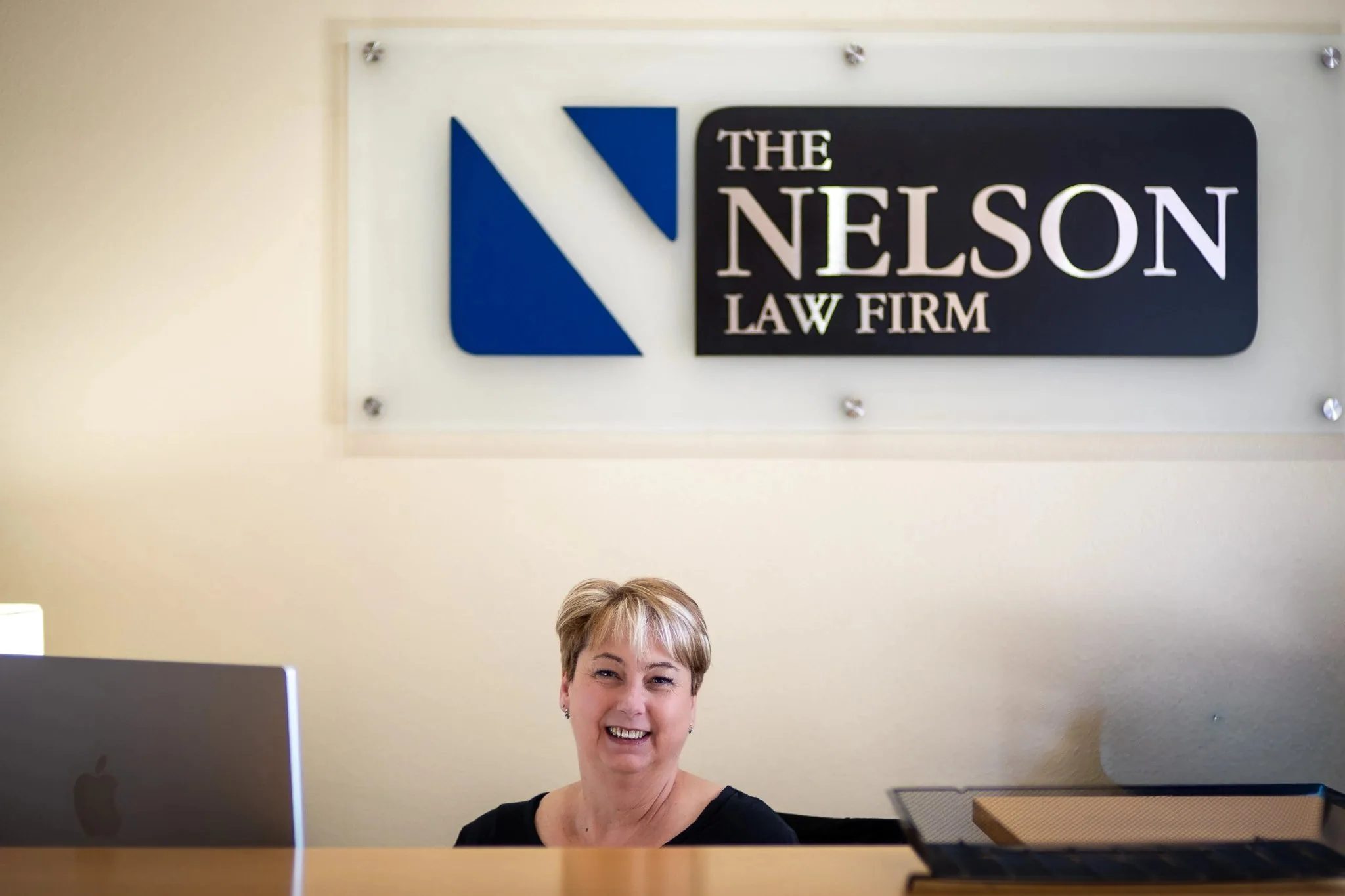 Dawn Schulte, Office Manager, smiles from behind the reception desk beneath the Nelson Law Firm sign.