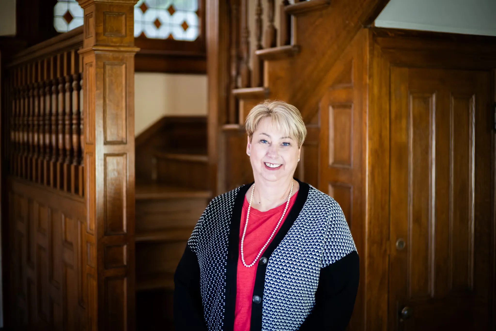 A Nelson Law Firm staff member in a red top and patterned cardigan smiling beside the oak staircase of the firm's Denver office