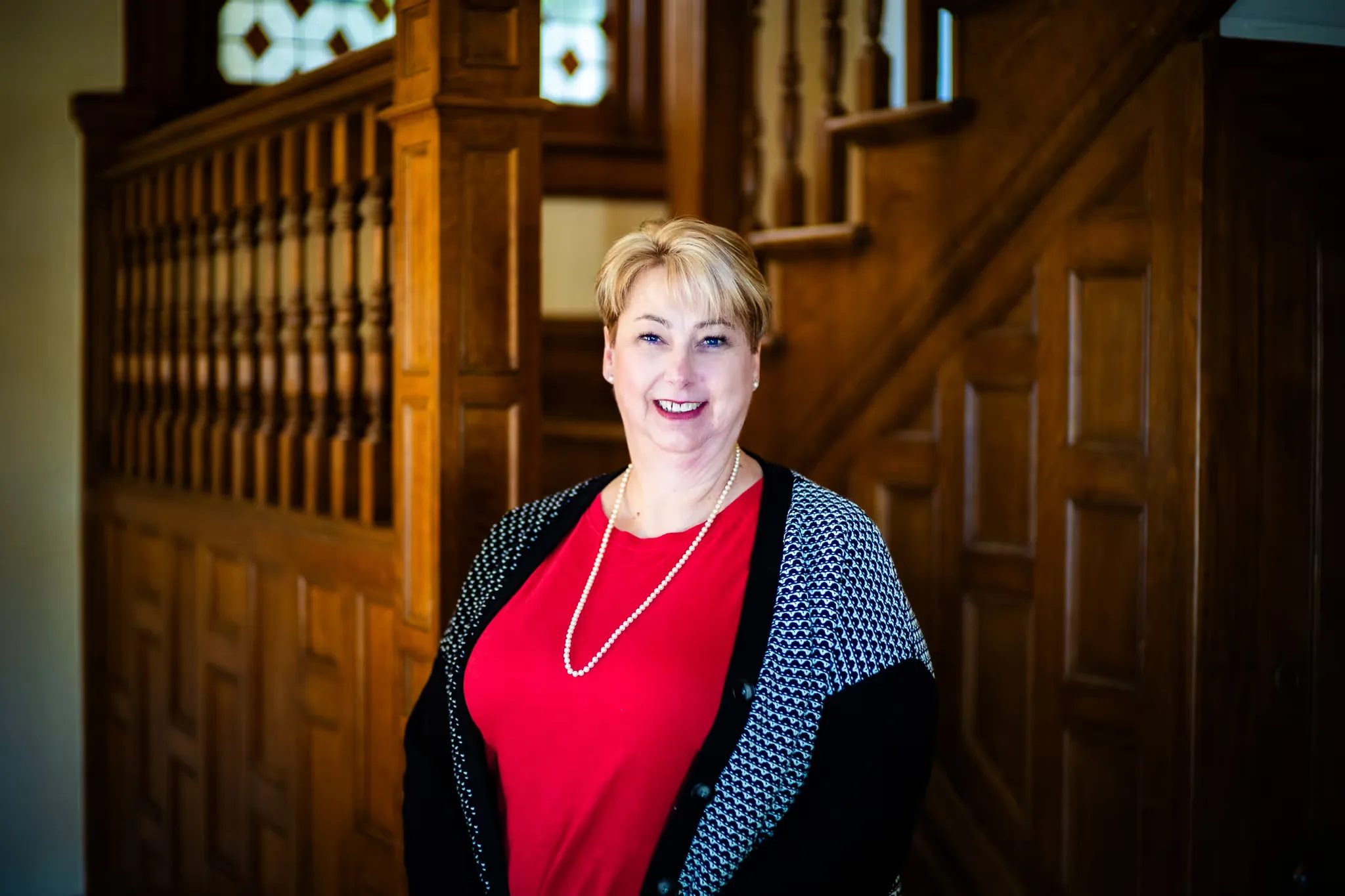 Close portrait of a Nelson Law Firm staff member in a red top and pearl necklace smiling at the firm's oak staircase in Denver