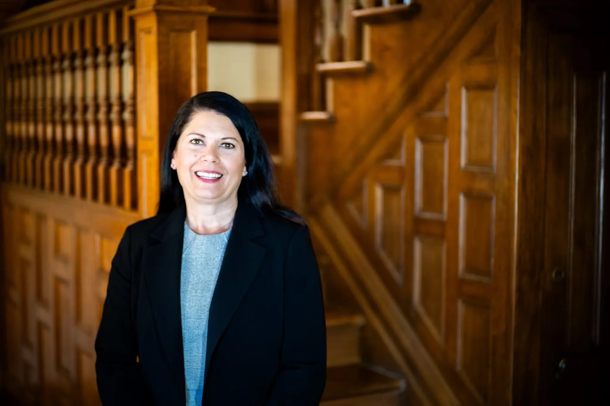 A Nelson Law Firm staff member in a black blazer and grey top smiling beside the oak staircase of the firm's Denver office