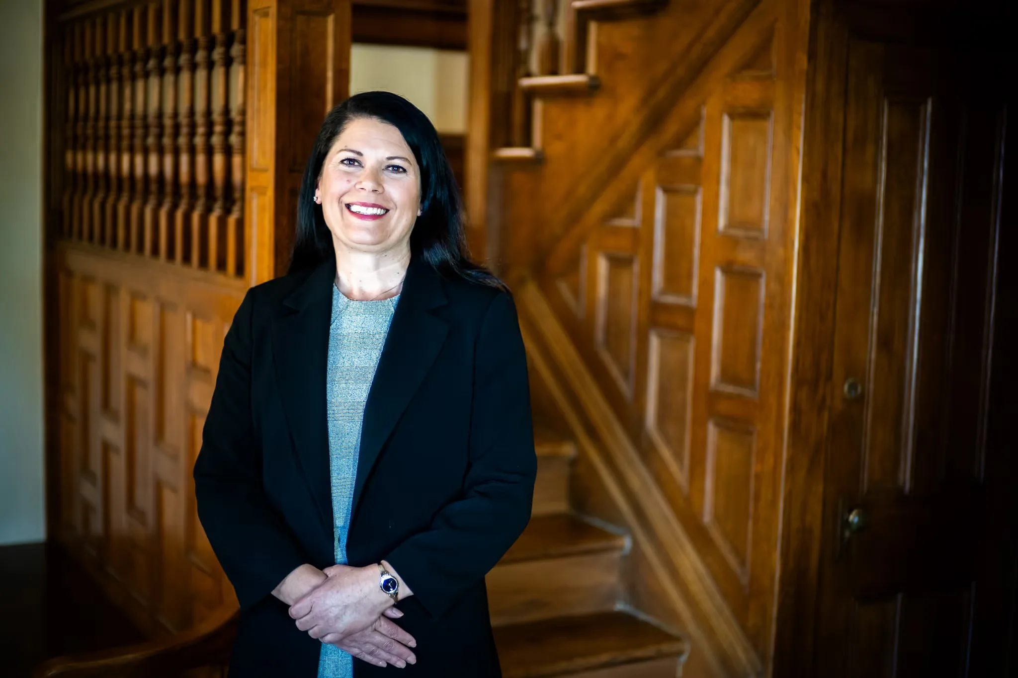 A Nelson Law Firm staff member in a black blazer and grey top with hands clasped at the firm's oak staircase in Denver