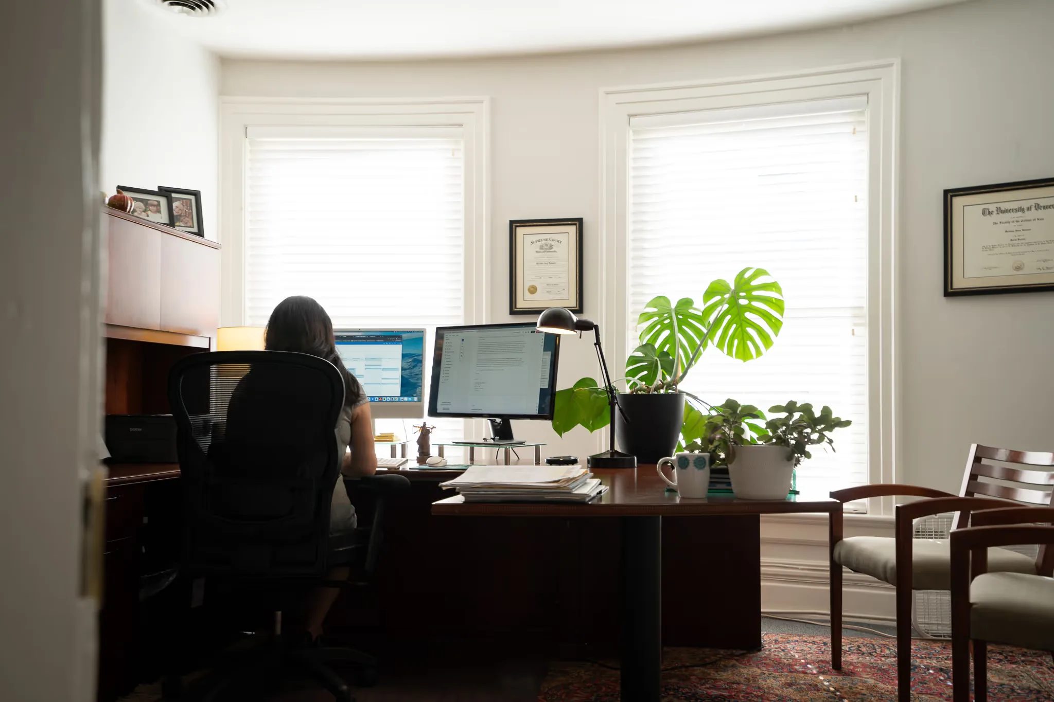 A bright corner office at Nelson Law Firm with a staff member at dual monitors, houseplants, framed diplomas and an area rug.