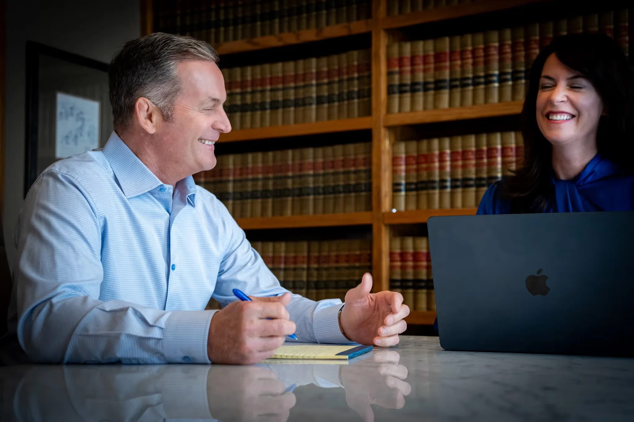 Mark W. Nelson and Colleen S. Nelson laughing across a conference table with a laptop and legal pad in the Nelson Law Firm library