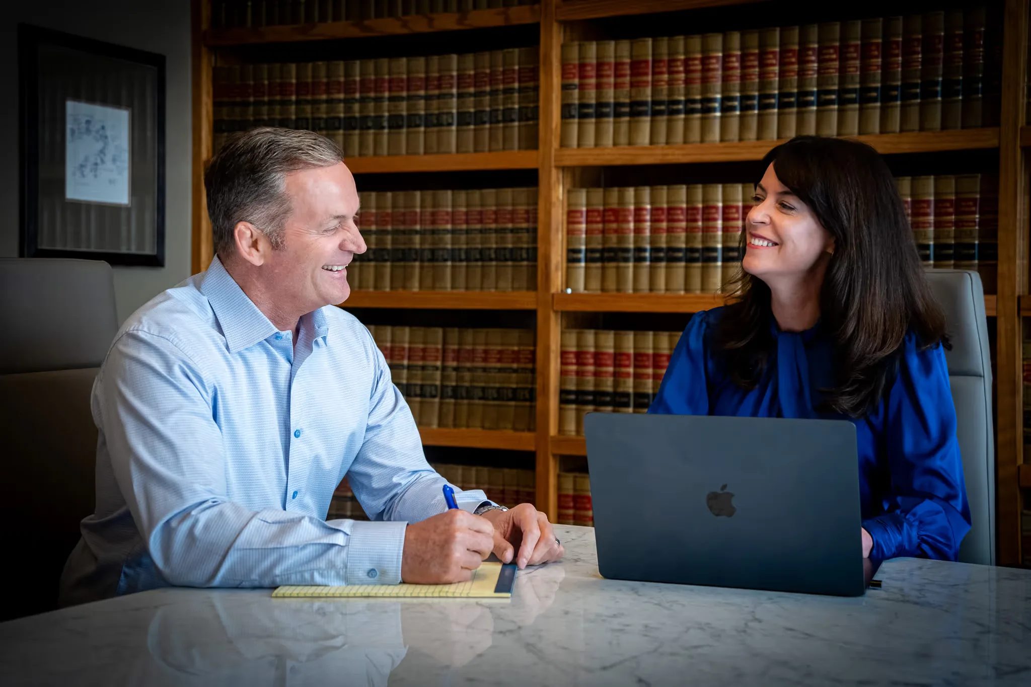 Mark W. Nelson and Colleen S. Nelson smiling at each other across a conference table with a laptop in the Nelson Law Firm library