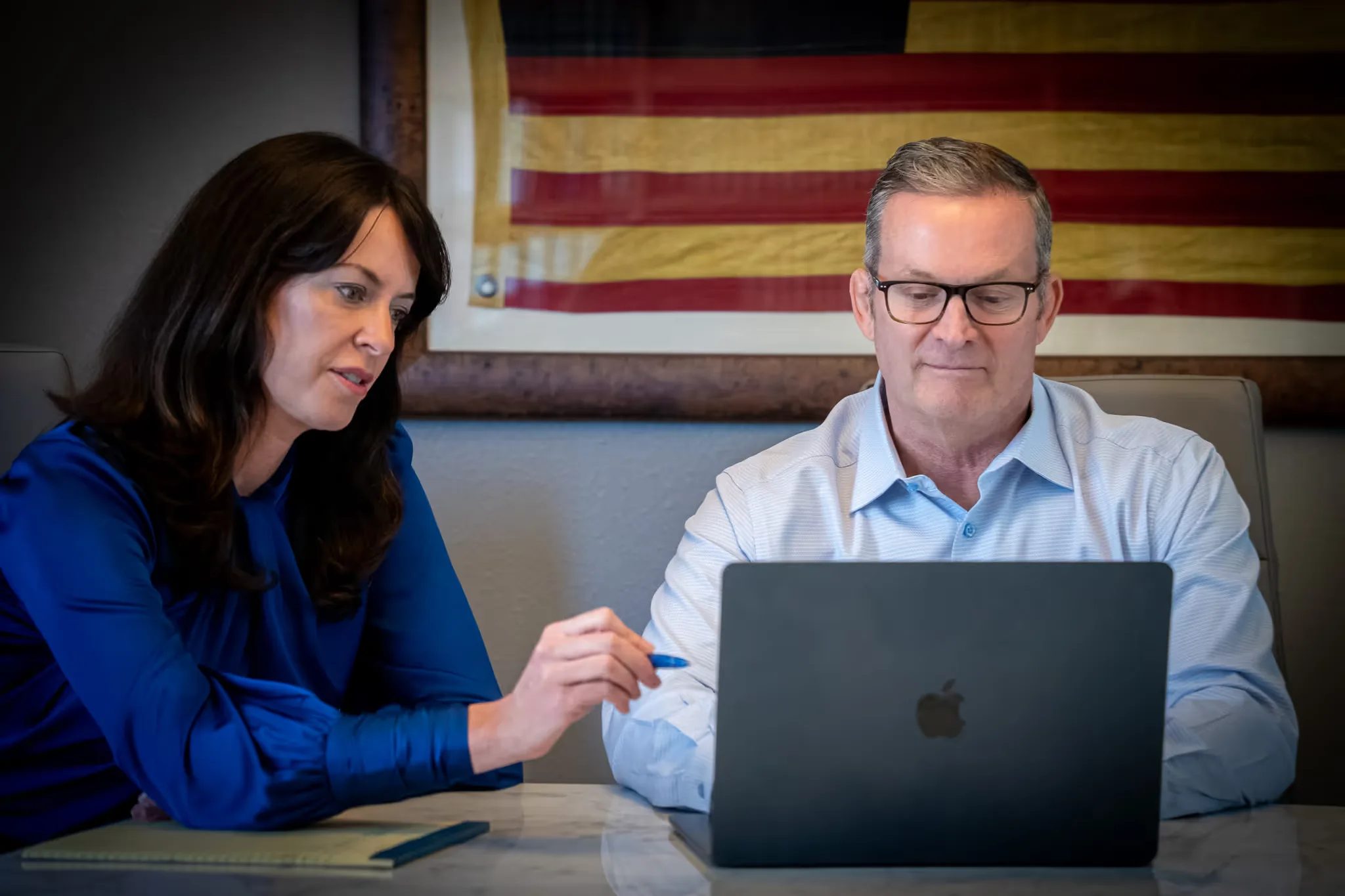 Colleen S. Nelson points at a laptop screen with a pen as Mark W. Nelson works, an antique American flag framed behind them