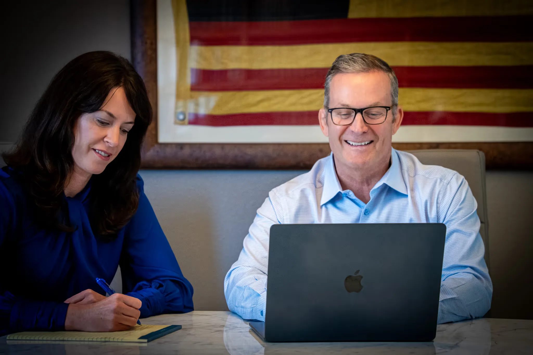 Colleen S. Nelson writing on a legal pad beside Mark W. Nelson at a laptop, a framed antique American flag behind them