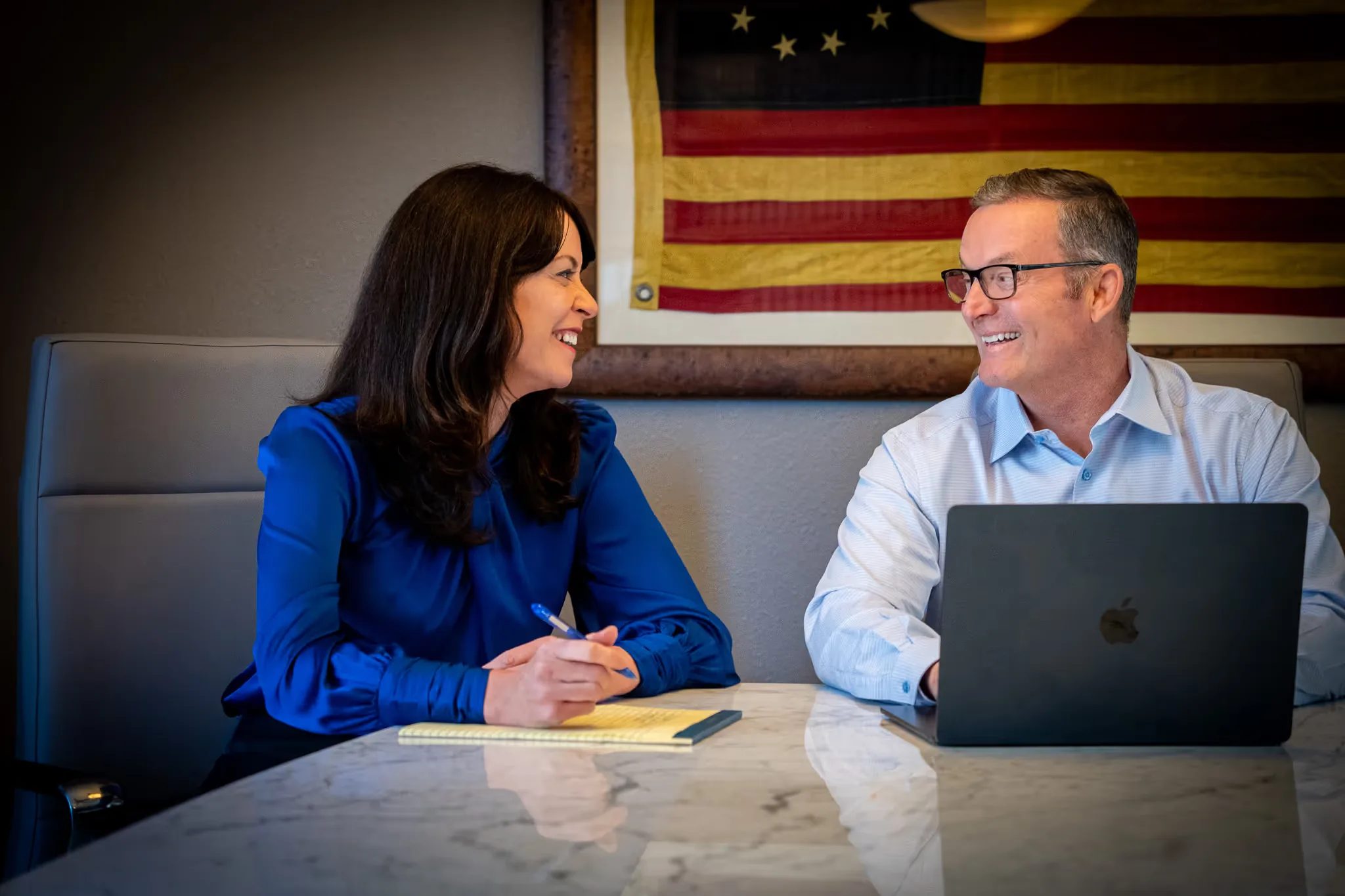 Mark W. Nelson and Colleen S. Nelson smiling at each other at a conference table under a framed antique American flag
