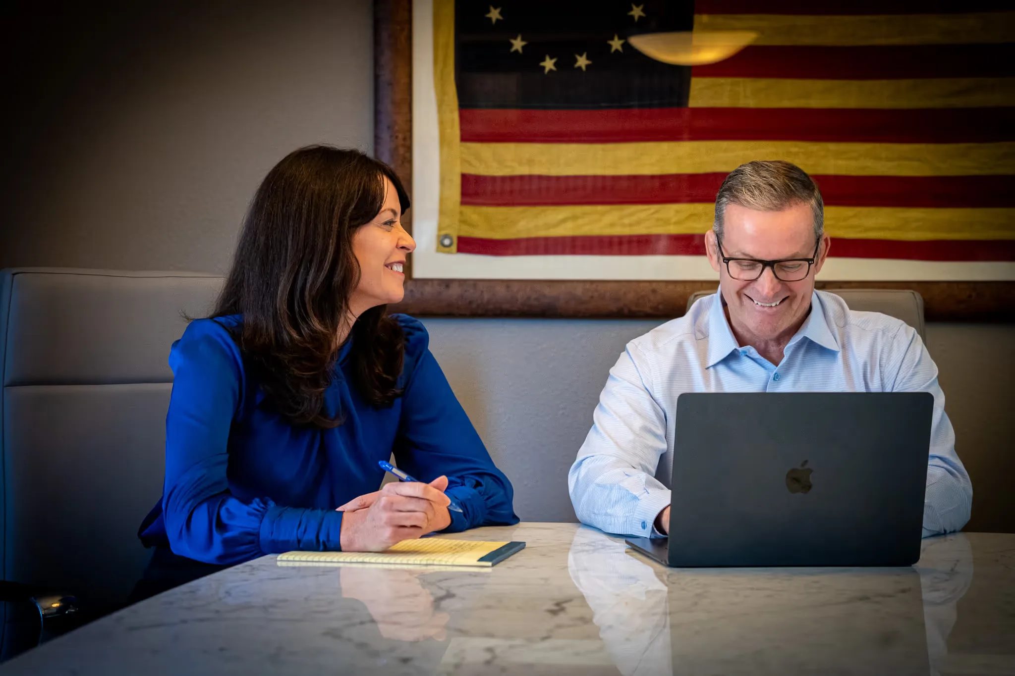 Colleen S. Nelson and Mark W. Nelson laughing together at a conference table with a laptop, antique American flag behind