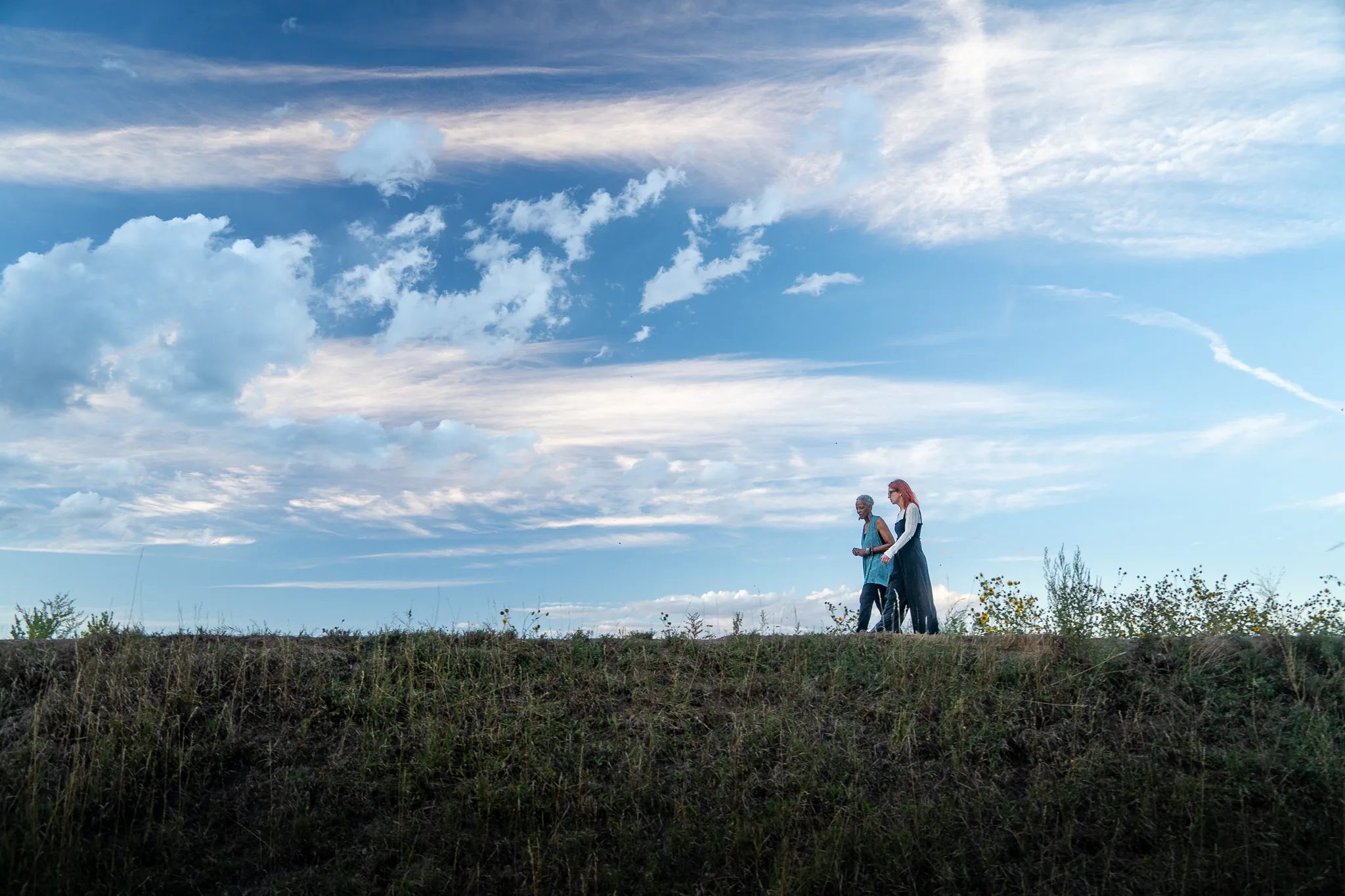 Norma Johnson and Amanda Cherry of Who Tells the Story walk along the crest of a grassy ridge beneath a wide cloud-streaked sky.