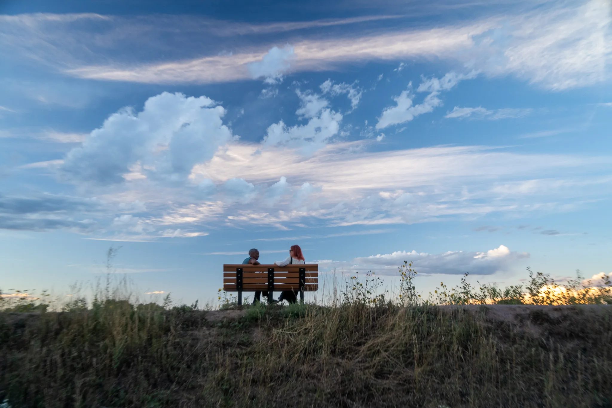 Who Tells the Story co-founders Norma Johnson and Amanda Cherry sit talking on a park bench atop a grassy ridge at sunset.