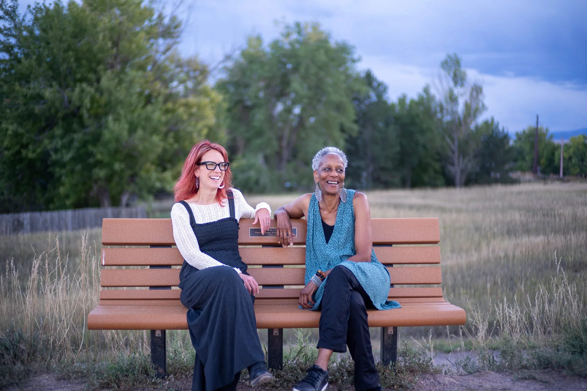 Who Tells the Story co-founders Amanda Cherry and Norma Johnson laugh together on a park bench in an open field.