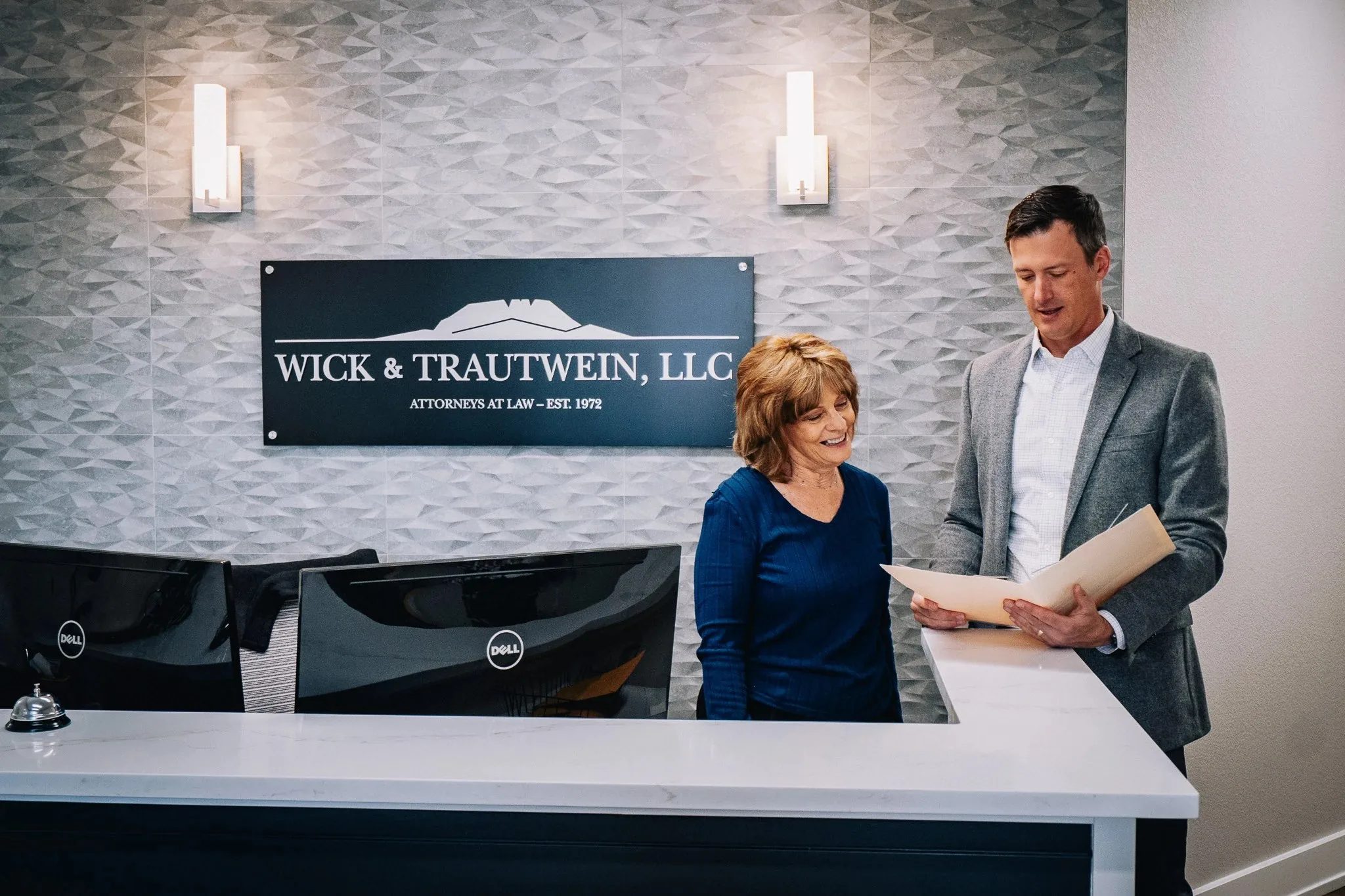 Two staff members at the Wick & Trautwein reception desk in Fort Collins, one reading from a folder beneath the firm's wall sign.