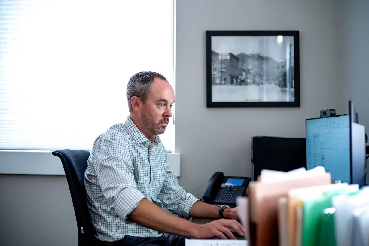 A staff member in a checked shirt works at a computer in a Wick & Trautwein office in Fort Collins, stacked file folders in the foreground.