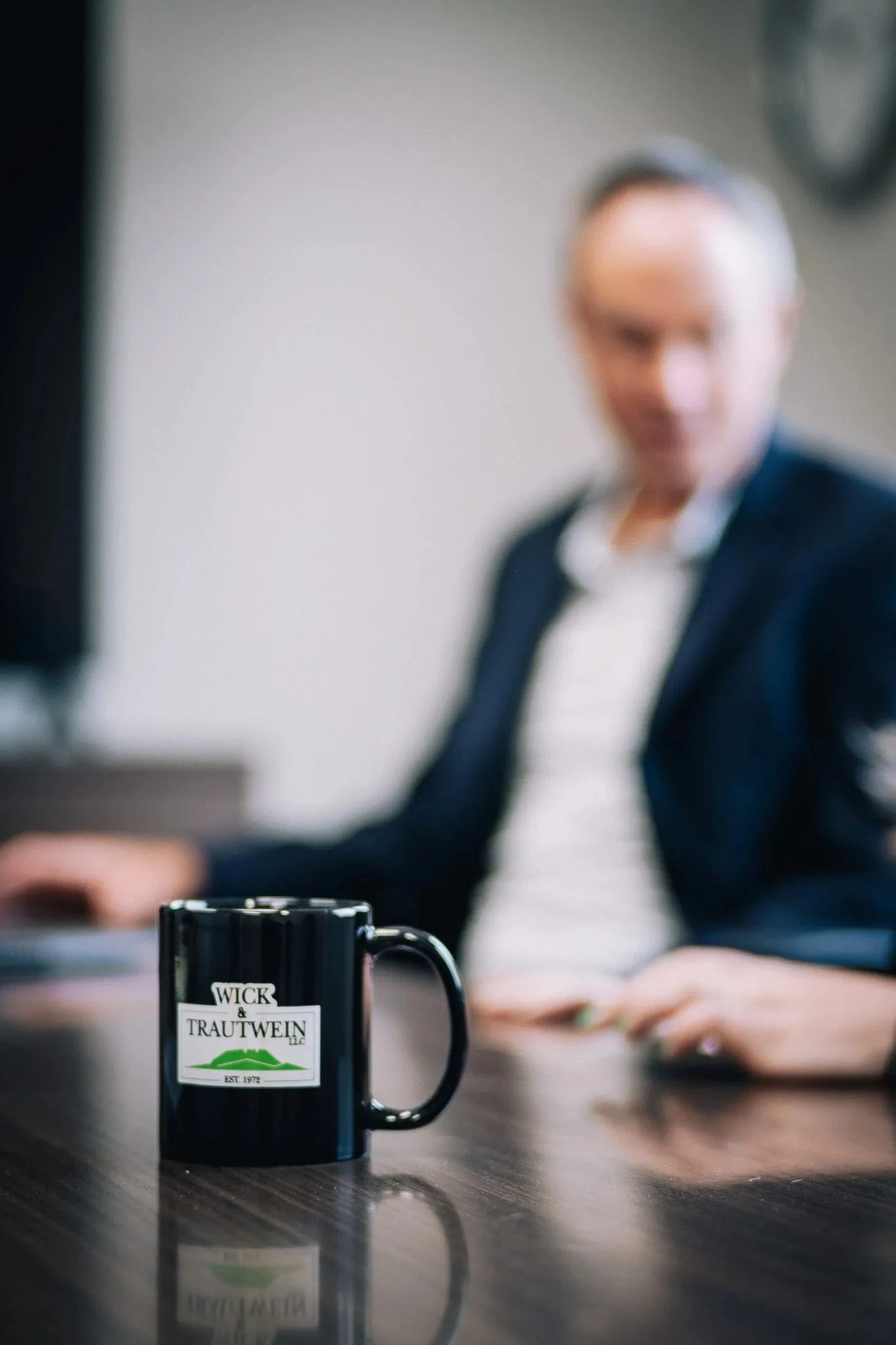 Black Wick & Trautwein logo mug on a conference table, with a staff member in a navy blazer seated out of focus behind it.