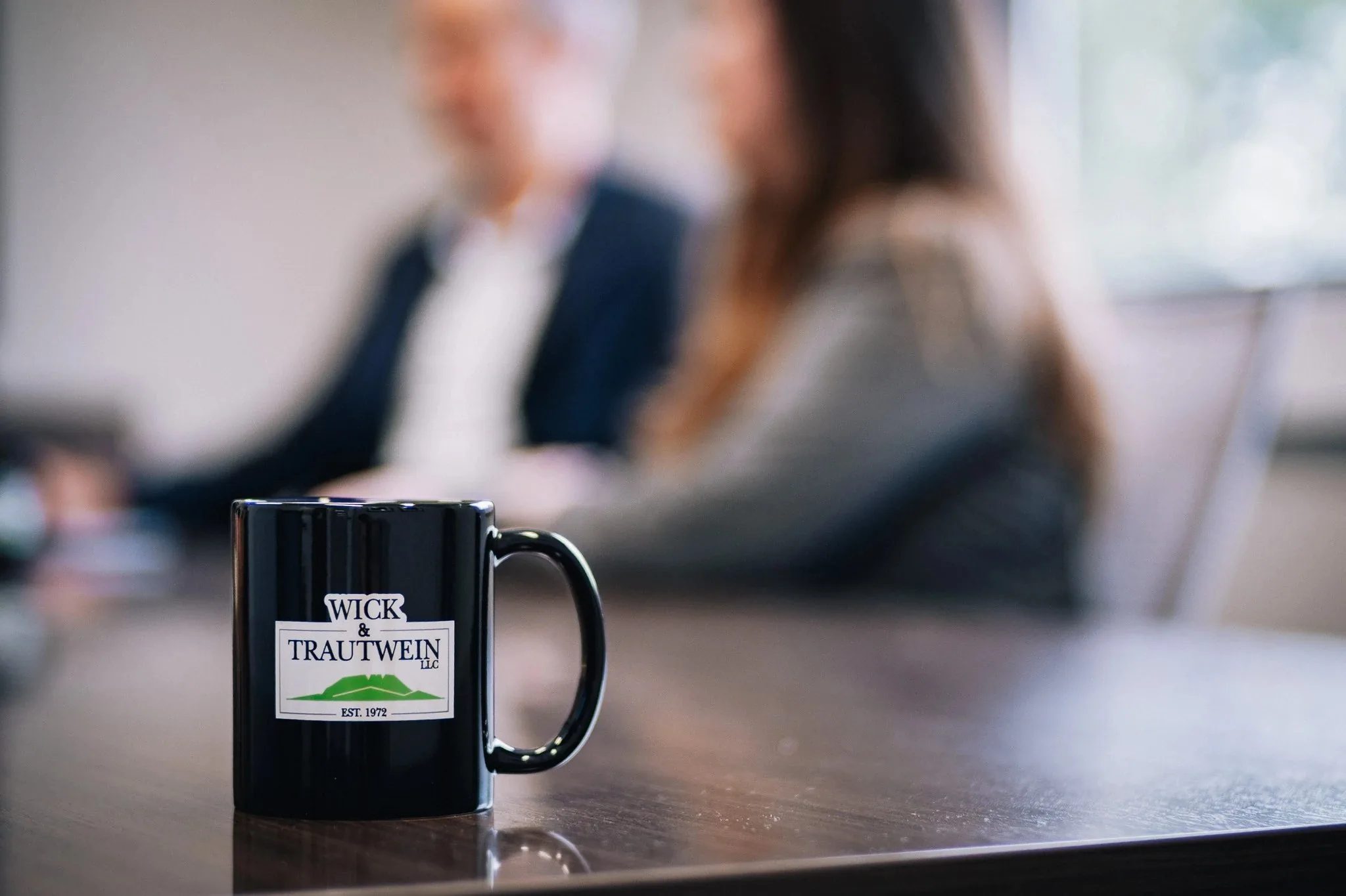 Wick & Trautwein logo mug in sharp focus on a conference table with two staff members blurred in the background.
