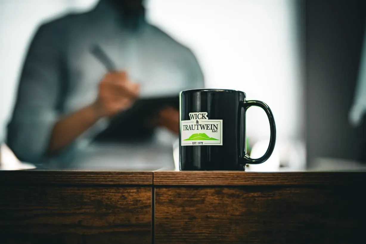 Black Wick & Trautwein logo mug on a wooden desk, with a staff member writing on a tablet out of focus behind it in Fort Collins.