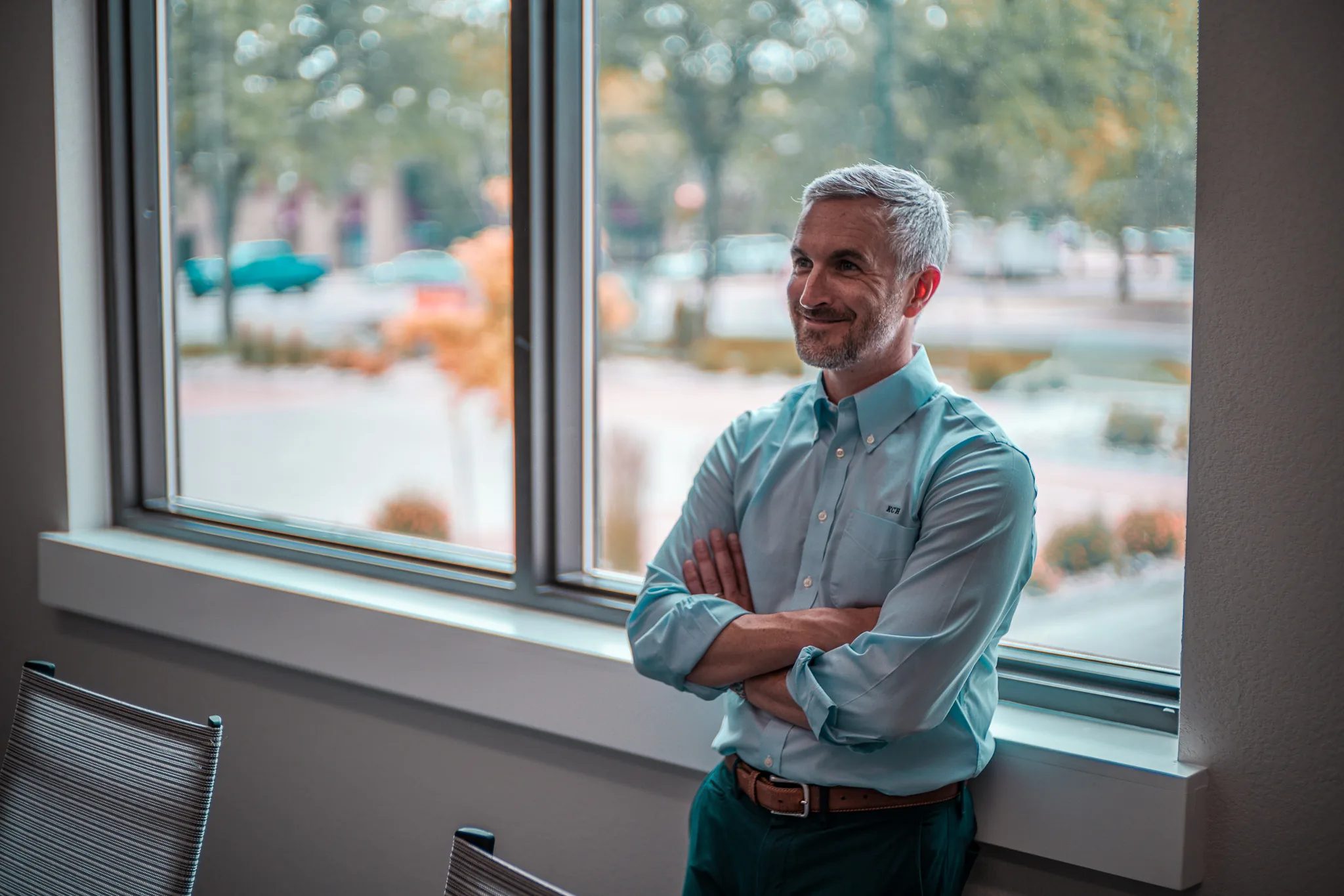 A smiling staff member with arms crossed stands beside an office window overlooking a tree-lined street at Wick & Trautwein in Fort Collins.