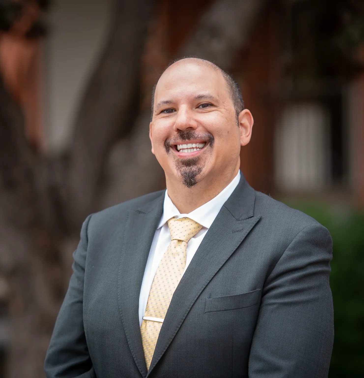 Outdoor headshot of a smiling person in a charcoal suit, white shirt and gold tie before a tree and brick building in Old Town Fort Collins.