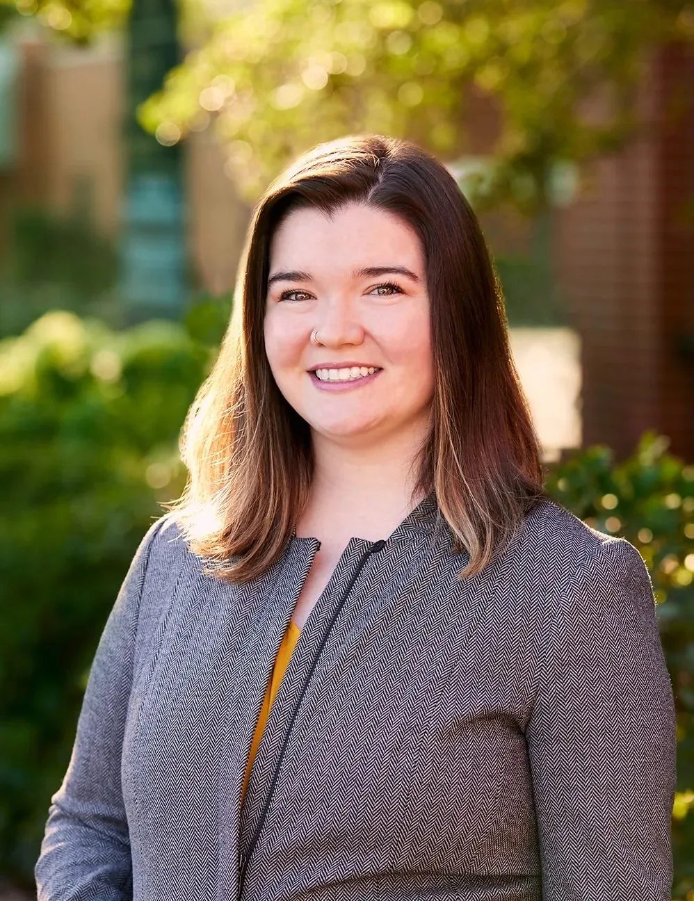 Cassie Williams, attorney at Wick & Trautwein, smiling in a gray herringbone blazer among sunlit trees and brick in Old Town Fort Collins.