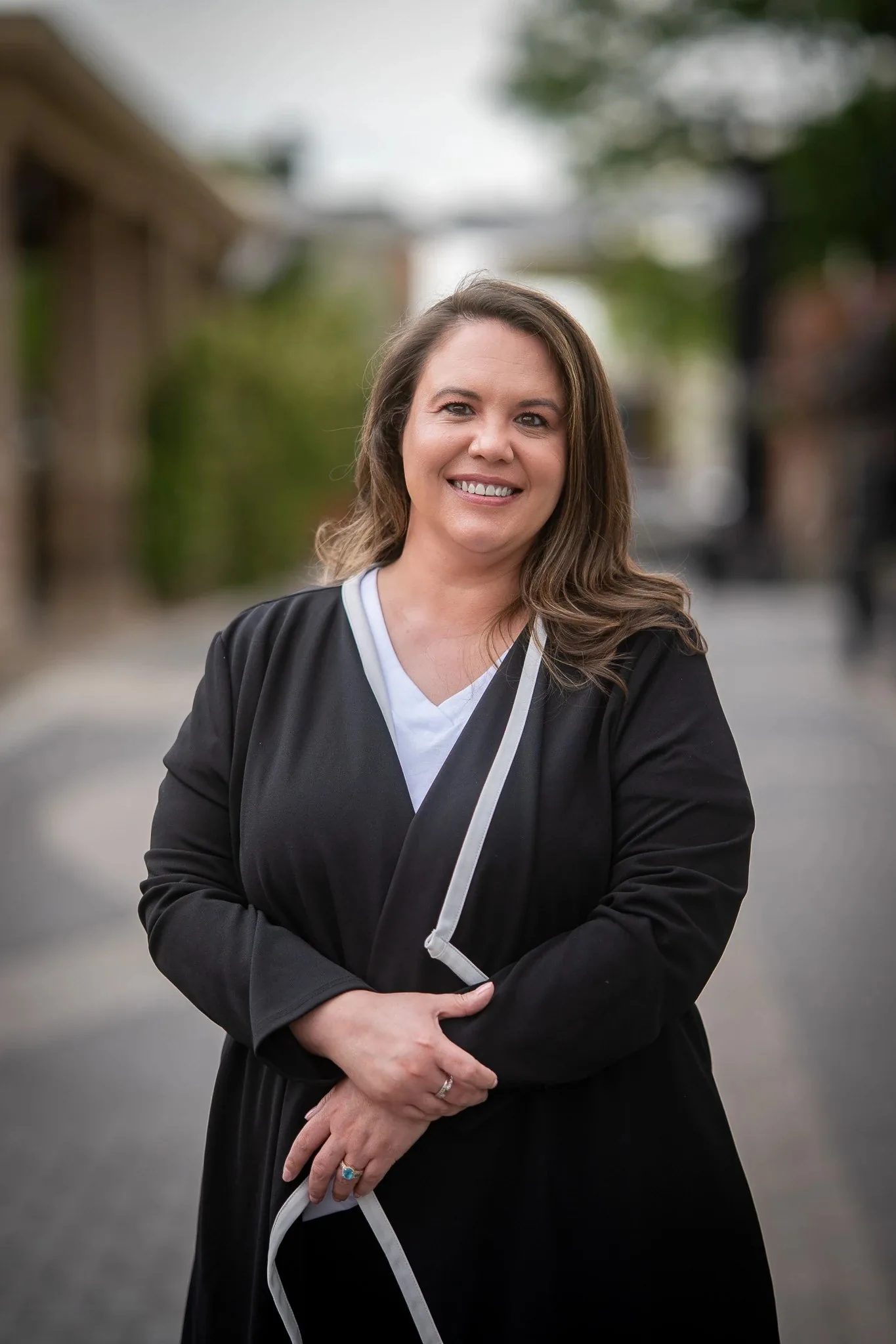 Outdoor headshot of a smiling person in a black cardigan with white trim, hands clasped, on a brick-paved street in Old Town Fort Collins.