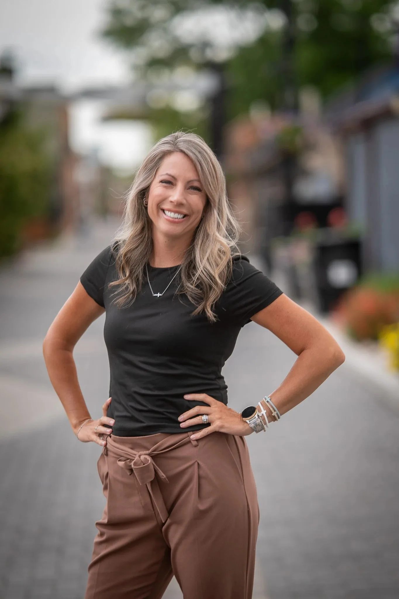 Outdoor headshot of a smiling person in a black t-shirt and brown trousers with hands on hips on a brick-paved street in Old Town Fort Collins.