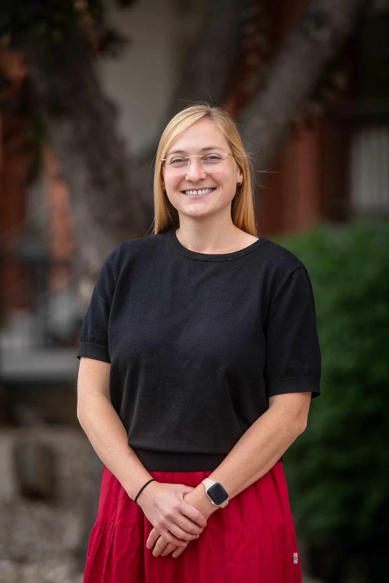 Outdoor headshot of a smiling person with glasses in a black top and red skirt, hands clasped, before a tree and brick wall in Old Town Fort Collins.