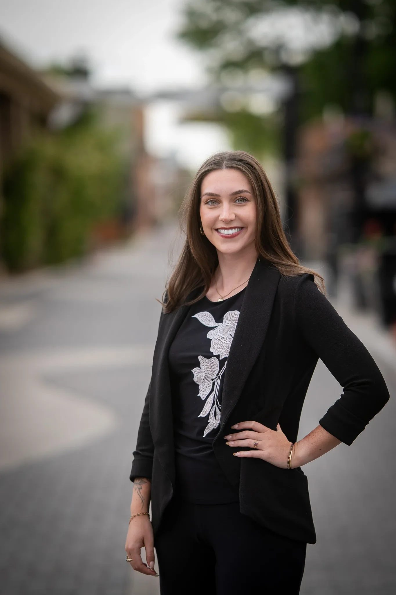 Outdoor headshot of a smiling person in a black blazer and floral-embroidered top, hand on hip, on a brick-paved street in Old Town Fort Collins.