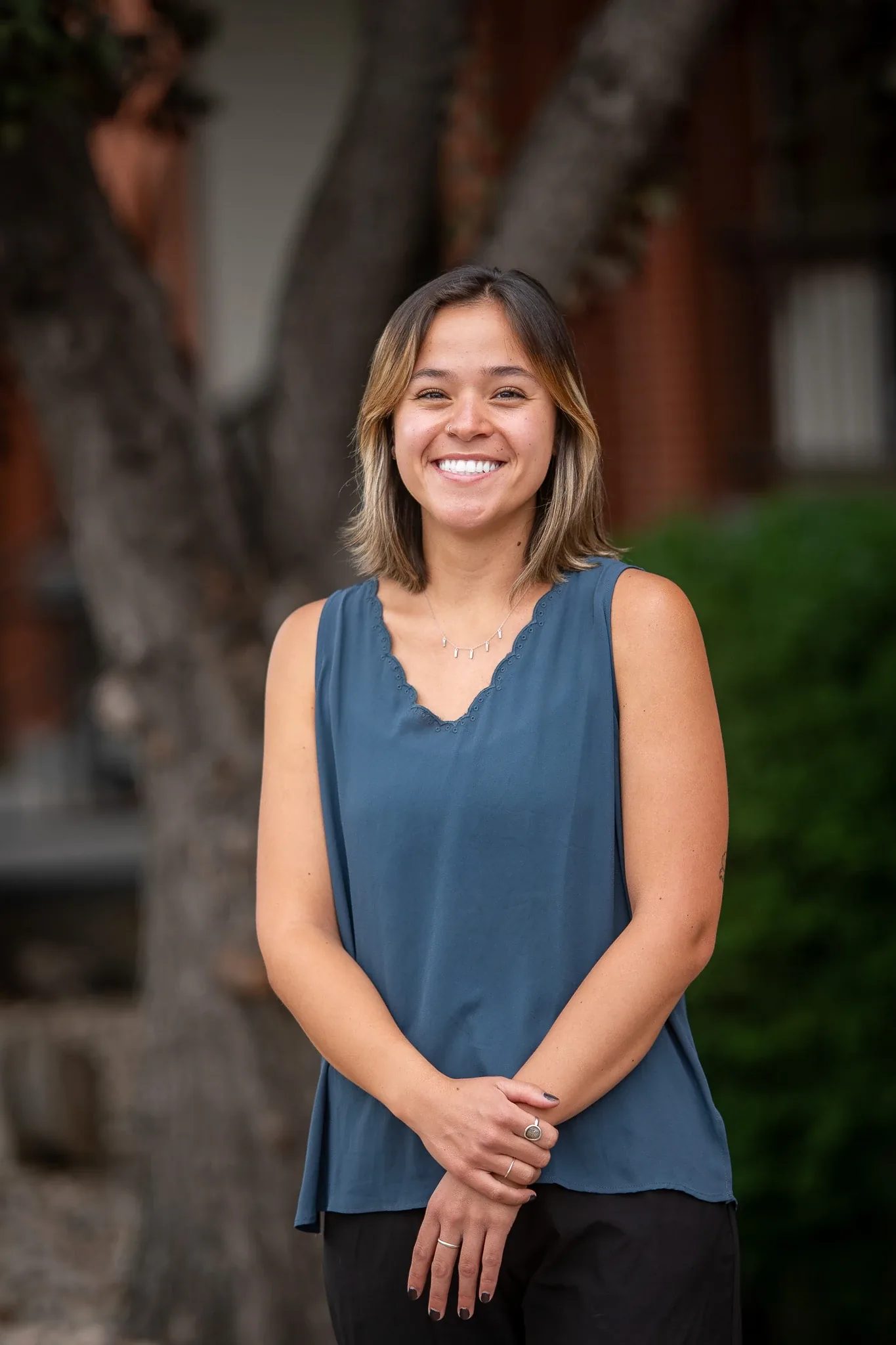 Outdoor headshot of a smiling person in a teal sleeveless top with hands clasped in front of a tree trunk and brick building in Old Town Fort Collins.