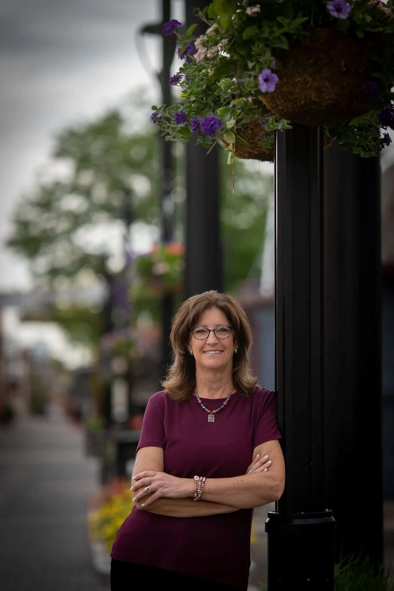 Outdoor headshot of a smiling person with glasses in a maroon top, arms crossed by a flower-basket lamp post in Old Town Fort Collins.