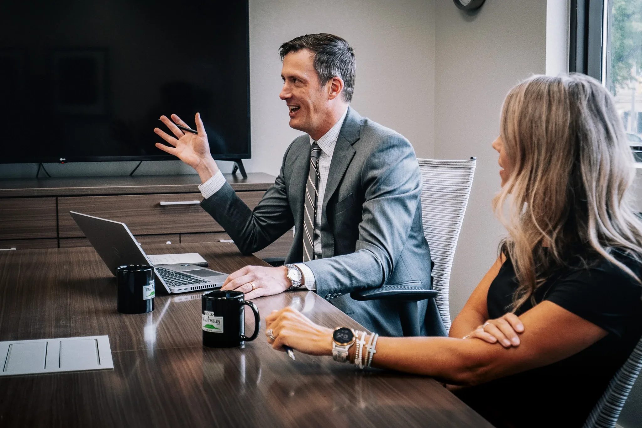 A person in a gray suit and striped tie gestures while speaking at a laptop across the Wick & Trautwein conference table in Fort Collins.