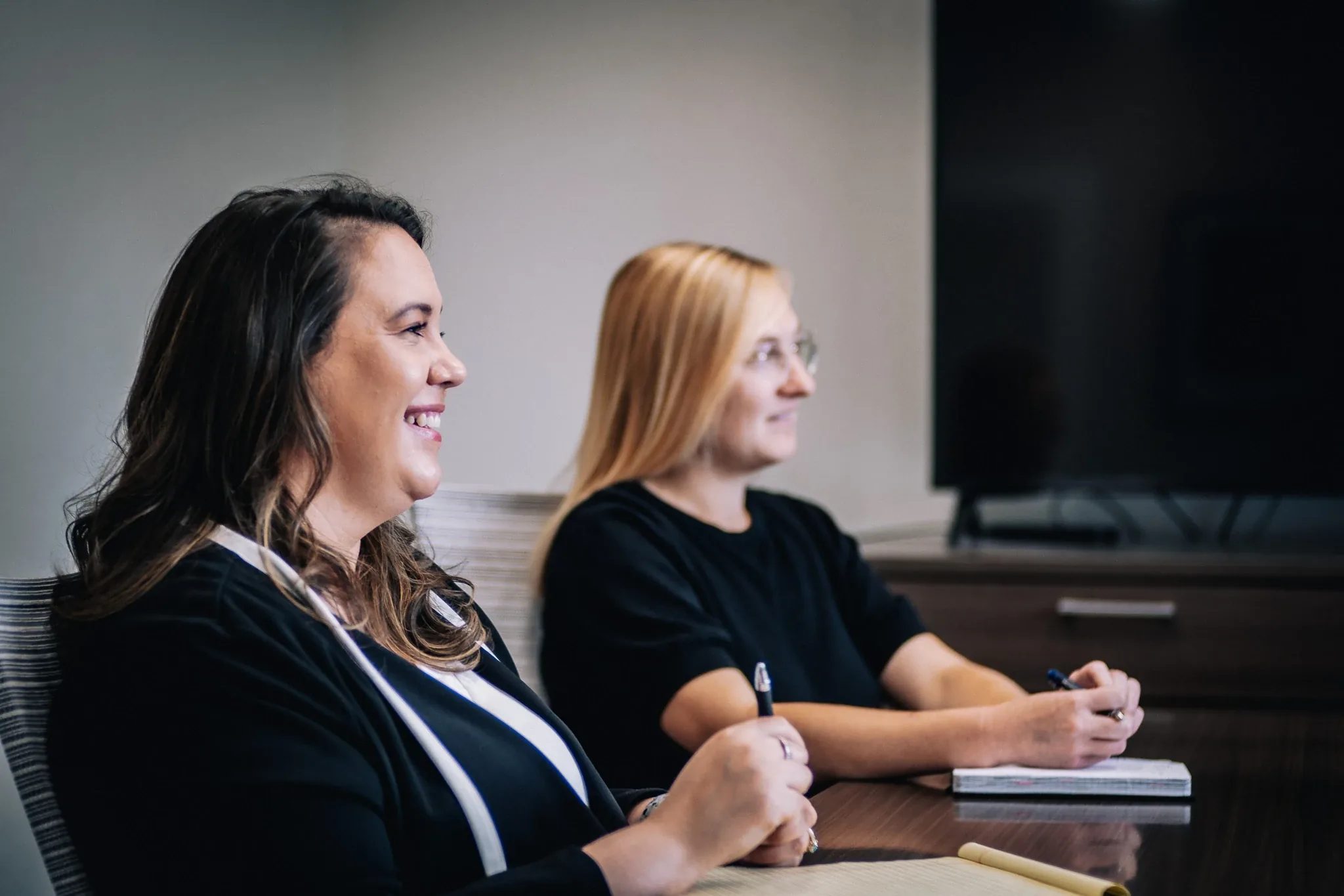 Two staff members seated at the Wick & Trautwein conference table in Fort Collins, smiling and holding pens as they listen.