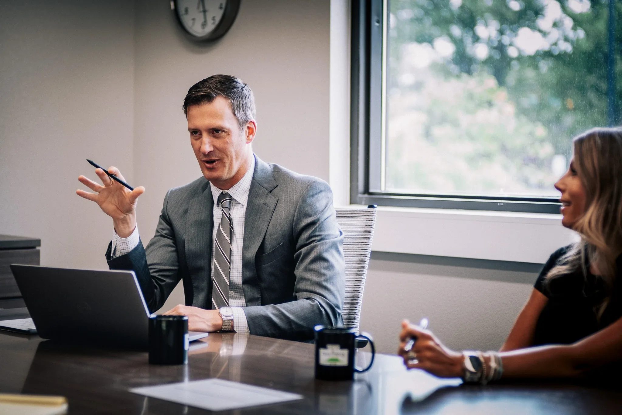 A person in a gray suit gestures with a pen while speaking at a laptop in the Wick & Trautwein conference room in Fort Collins.