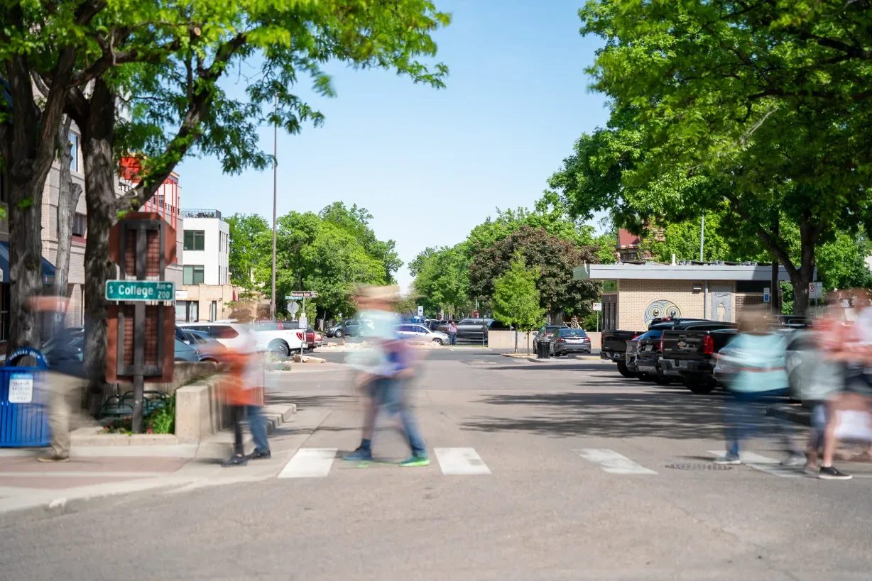 Pedestrians blur through a crosswalk at South College Avenue in Old Town Fort Collins on a sunny day, framed by leafy trees.