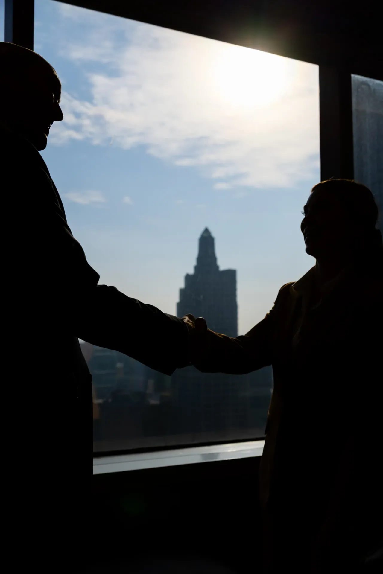 Two people shake hands, silhouetted against a bright office window overlooking a city.