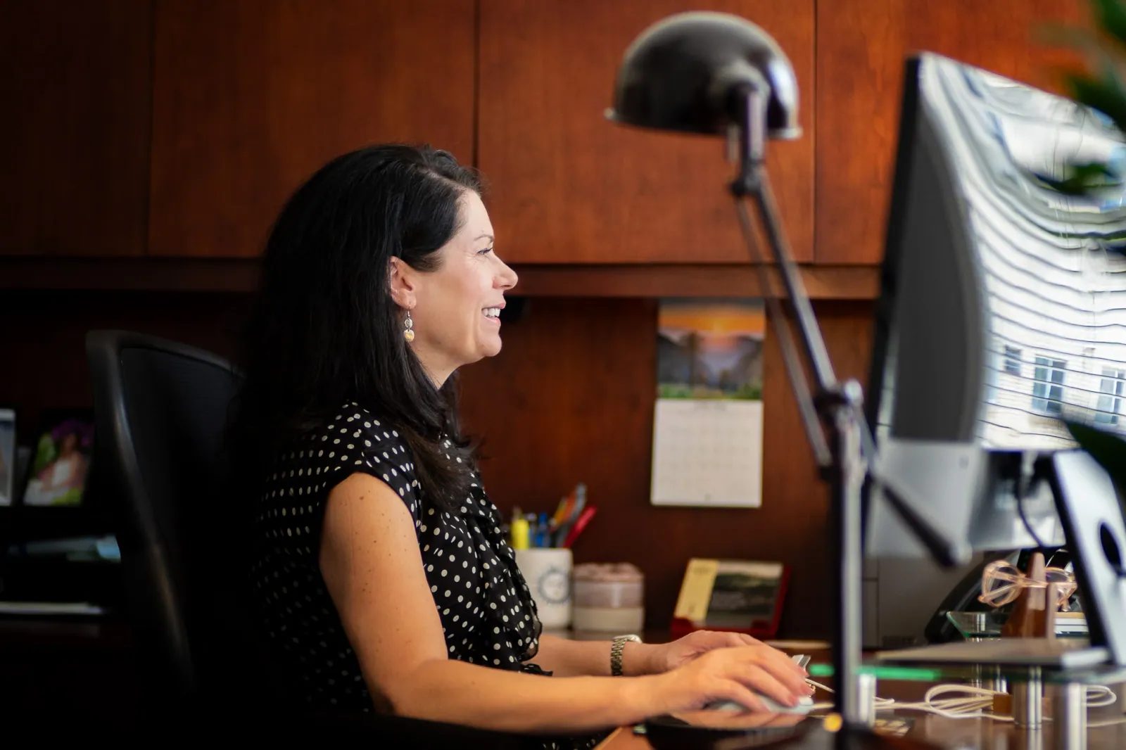 A woman works at her desktop computer in a warmly lit office.