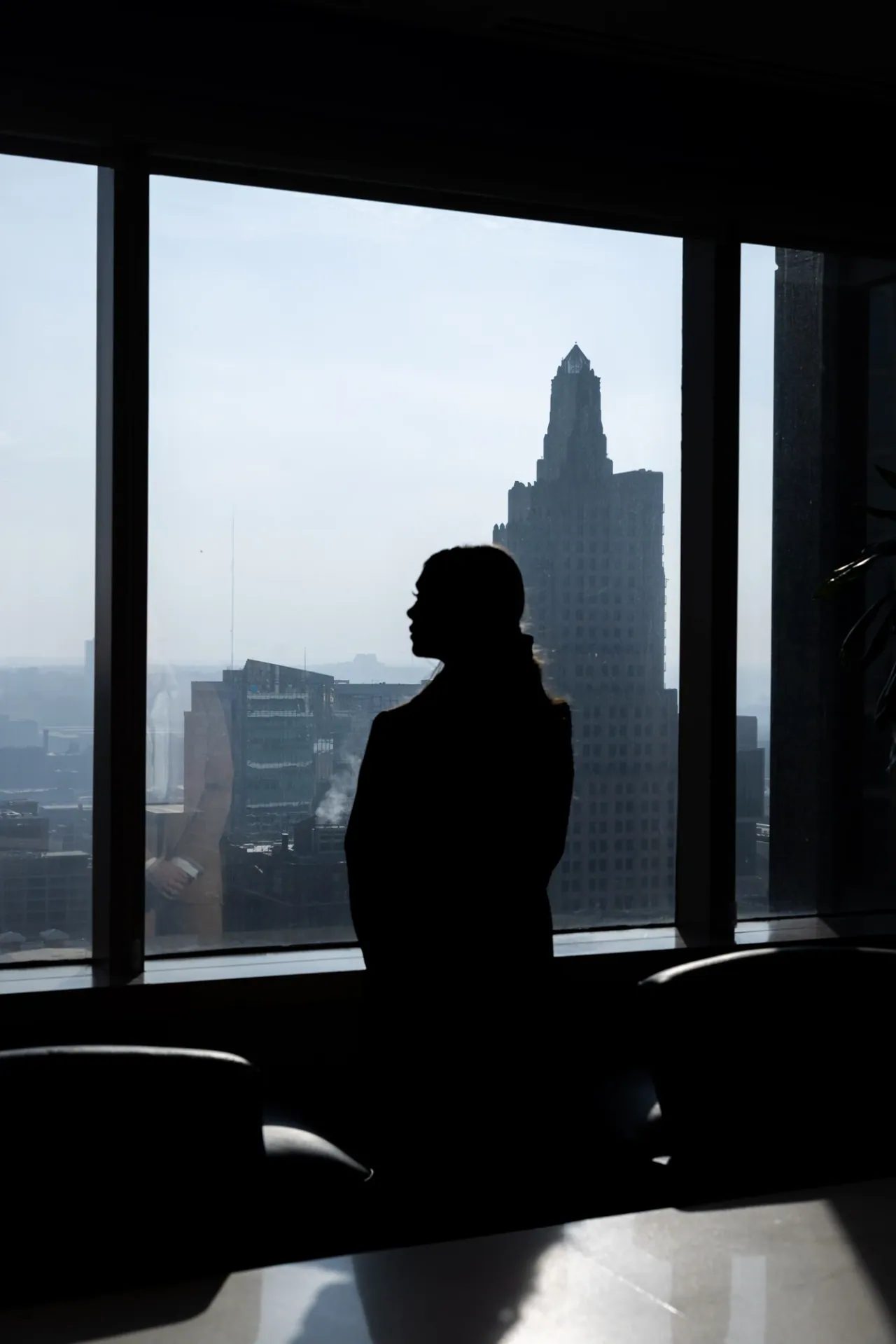 A person stands at a floor to ceiling window looking out over a hazy city skyline.