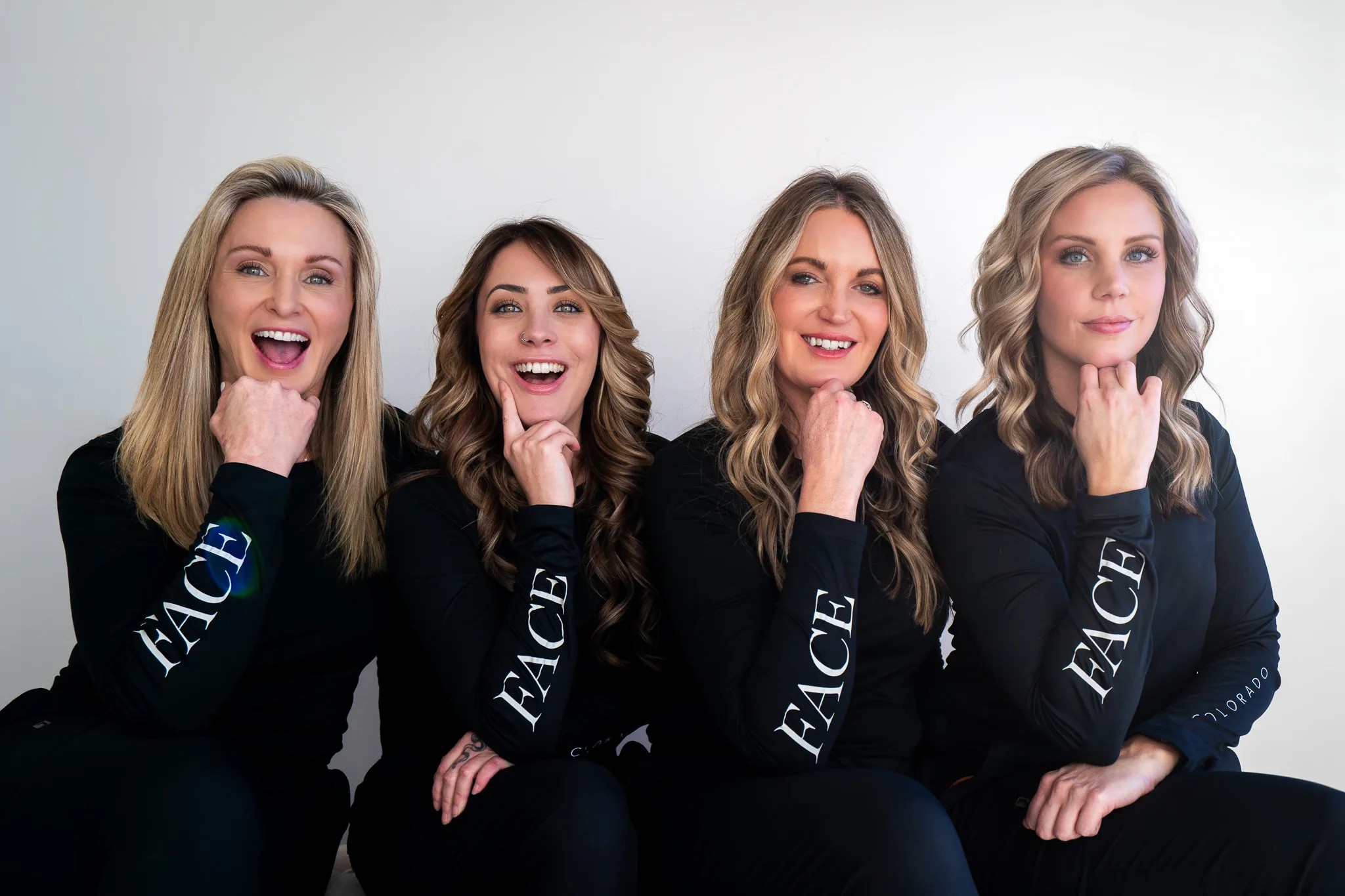 Four FACE Colorado team members in matching black FACE long-sleeve tops sit in a row, chins on their hands, laughing at the camera.