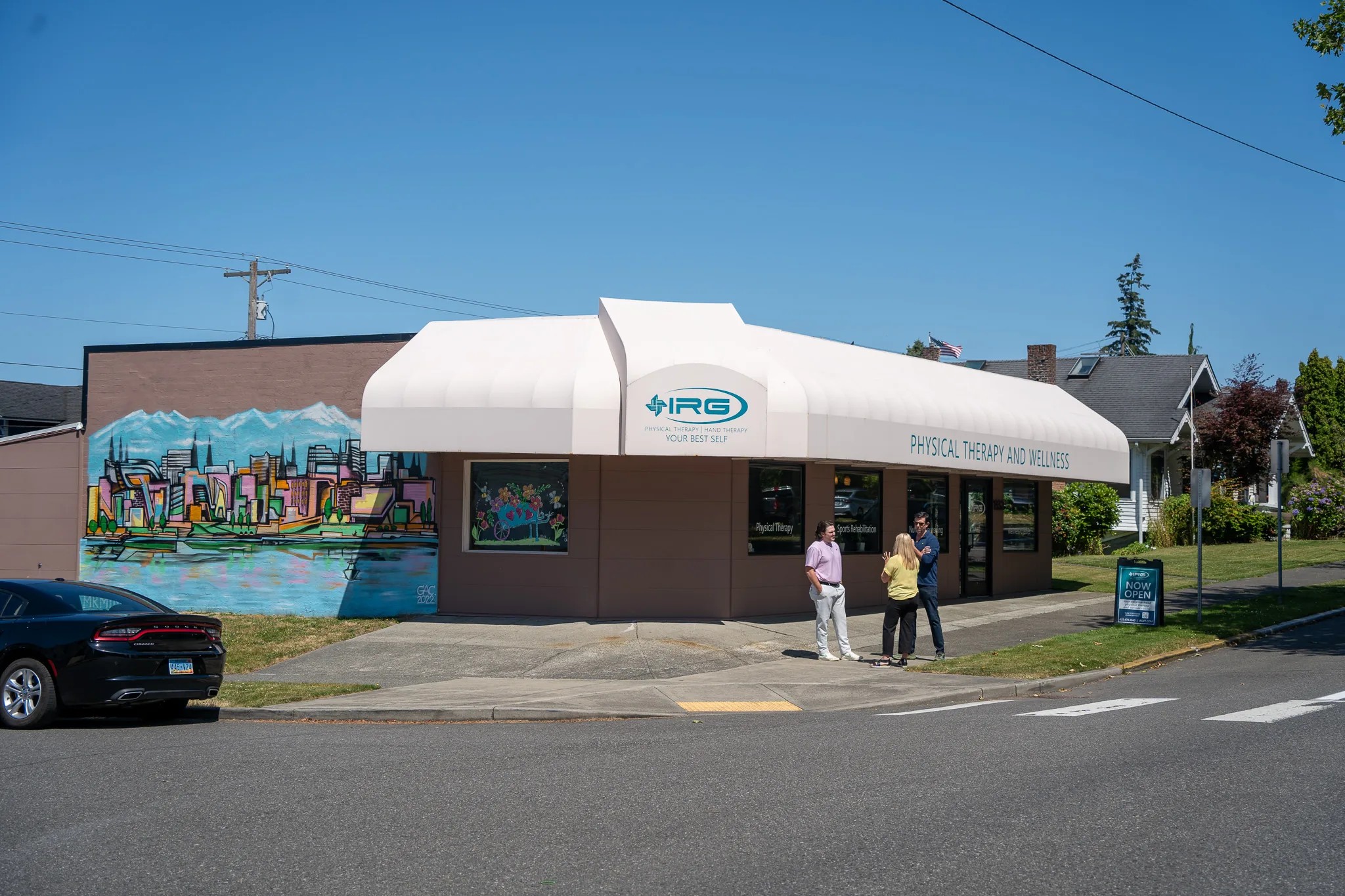 A corner clinic with a white IRG Physical Therapy and Wellness awning beside a painted city mural, three people talking on the sidewalk.
