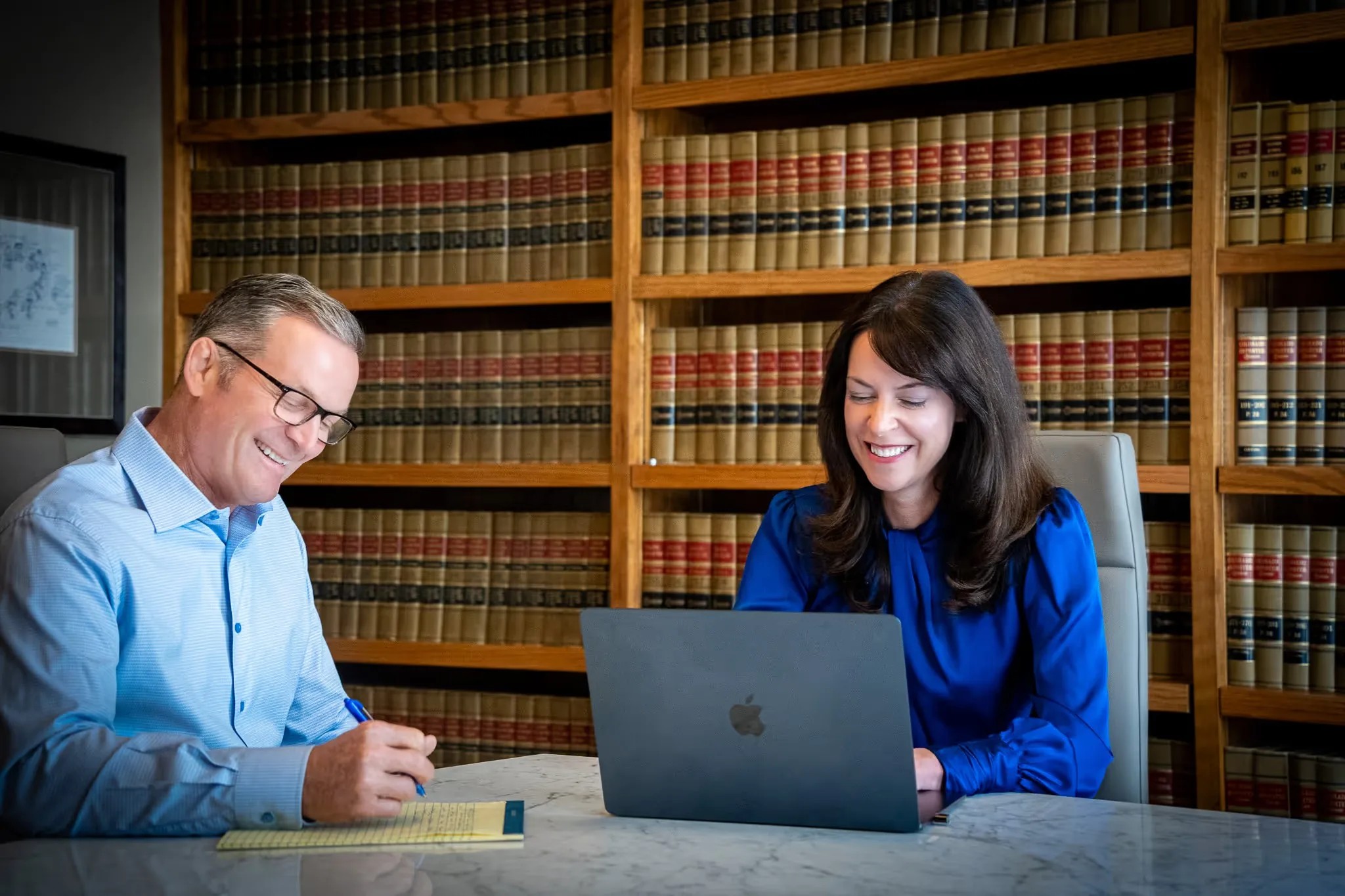 Two members of the Nelson Law Firm team work together at a marble table in the law library, one on a laptop and one taking notes.