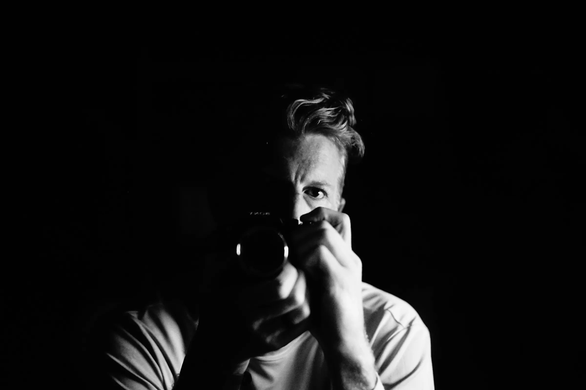 Black-and-white self-portrait of photographer Cody Strate, half his face lit, holding a camera to his eye against a black background.